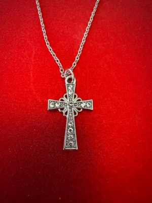 Kedja med glittrigt korshänge - Säljer ett halsband med ett silverfärgat korshänge dekorerat med gnistrande stenar. Korset har en detaljrik design med små blommönster runt mitten och sitter på en tunn kedja. Perfekt accessoar för dig som gillar statement-smycken.