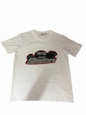 Vit Trapstar London Shooters t-shirt - Säljer en clean vit t-shirt från Trapstar med stort broderat 'London Shooters'-motiv och panter på bröstet. Klassisk rund hals och korta ärmar. Skön bomullskvalitet och streetwear-vibe, perfekt till jeans eller shorts.
