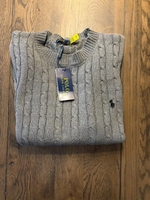 Grå kabelstickad tröja Polo Ralph Lauren - Snygg grå kabelstickad tröja från Polo Ralph Lauren med klassisk rund hals och broderad logga på bröstet. Tröjan har långa ärmar och ribbade muddar vid ärmslut och nederkant. Perfekt för lager-på-lager under kyliga dagar.