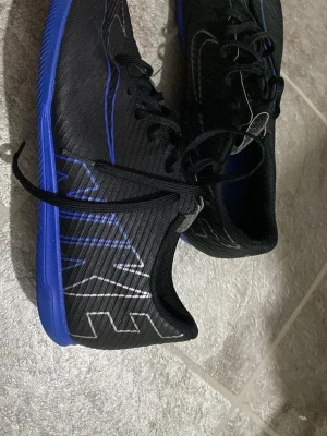  Nike futsalskor - Säljer ett par svarta Nike futsalskor med blå sula och blå detaljer. Skorna har snörning och är tillverkade i syntetmaterial. Stora Nike-loggor i vitt och blått på sidorna ger en sportig look. Perfekta för dig som vill sticka ut på planen.