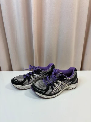 Lila och svarta Asics sneakers - Säljer ett par Asics sneakers i svart, vitt och lila med mesh-överdel och klassiska Asics-loggan på sidan. Skorna har snörning, platt sula och vadderad insida för extra komfort. Perfekta för träning eller vardagsbruk.