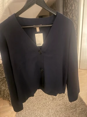 Mörkblå kofta från H&M, S - Mysig mörkblå kofta från H&M i storlek S. Koftan har v-ringning, två knappar framtill och långa ärmar. Perfekt att slänga över en t-shirt eller linne för en chill look. Materialet känns mjukt och skönt mot huden.