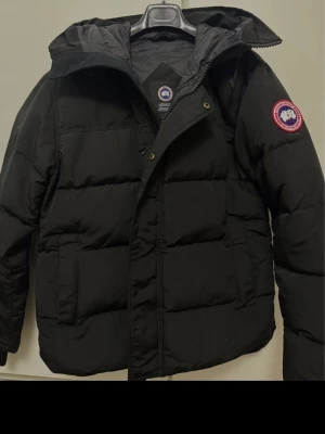 Svart dunjacka från Canada Goose - Säljer en svart, klassisk dunjacka från Canada Goose i storlek L. Jackan har huva, dragkedja och tryckknappar framtill samt den ikoniska loggan på ärmen. Fylld med dun och fjädrar för maxad värme, perfekt för kalla dagar. Snygg, enkel och riktigt varm.