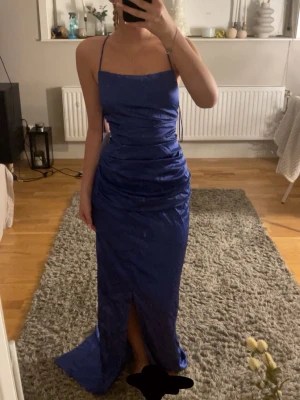 Blå balgklänning med öppen rygg - Säljer en snygg blå långklänning med smala axelband. Klänningen har öppen rygg med korsade band och knytning, samt en slits framtill, då klänningen var alldeles för lång för mig så sitter slitsen nog lite långt ner så kolla på sista bilden för att se hur jag TROR den ska sitta!💞