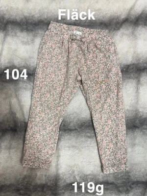 Blommiga byxor med snörning, stl 104 - Söta byxor med blommigt mönster i rosa, vitt och grönt. Byxorna har snörning i midjan. Perfekta för dig som gillar en chill och lekfull stil.🌸 Fläck. Storlek 104