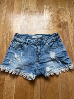Blå jeansshorts med spets från Amisu - Säljer ett par ljusblå jeansshorts från Amisu med snygga slitningar och vita spetsdetaljer längs bensluten. Klassisk femficksmodell med knapp och dragkedja framtill. Perfekta för sommardagar och ger en söt touch till din outfit.