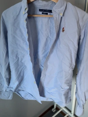 Ljusblå skjorta från Ralph Lauren - Säljer en ljusblå skjorta från Ralph Lauren i slim fit-modell. Skjortan har klassisk krage, knappar framtill och den ikoniska broderade loggan på bröstet. Perfekt för dig som gillar stilrena och tidlösa plagg.