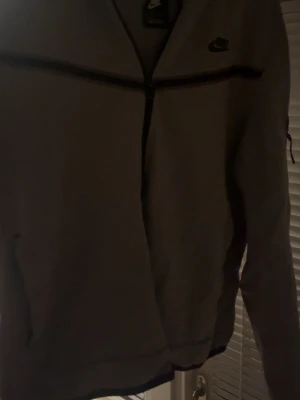 Grå Nike zip hoodie med svarta detaljer - Säljer en grå hoodie från Nike med hel dragkedja och svarta detaljer längs bröstet och ärmen. Tröjan har lång ärm och en ficka med dragkedja på ena ärmen. Perfekt för dig som gillar sportig och clean stil.