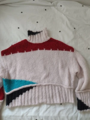 Stickad tröja i pastell och rött - Säljer en stickad tröjai ljusrosa med röda, svarta och turkosa detaljer. Tröjan har hög krage, ribbade muddar och en croppad passform. Perfekt för dig som gillar färg och unika plagg. Supermysig och varm för kyliga dagar. 