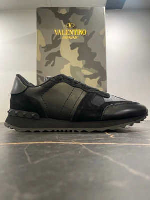 Valentino Garavani Rockrunner sneakers - Svarta Valentino Garavani Rockrunner sneakers med coola nitar på hälen och diskret camo-mönster. Skorna har en mix av skinn, mocka och textil, samt snörning och platt sula. Perfekta för dig som vill ha en lyxig och edgy vibe till din outfit.