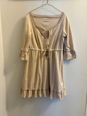 Beige Odd Molly klänning med volang - Supersöt beige klänning från Odd Molly med volangkant nertill och vid ärmslut. Klänningen har knytband i midjan och vid halsen, spetsdetaljer runt urringningen och en broderad fjäril på bröstet. Perfekt för dig som gillar romantisk och avslappnad stil.