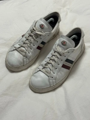 Vita Moncler sneakers med röd och marinblå detalj - Säljer ett par vita sneakers från Moncler i skinn med klassiska röd och marinblå ränder på sidorna och Moncler-logga på plösen. Skorna har snörning och en platt sula, perfekt för dig som gillar stilrena och sportiga vibes.