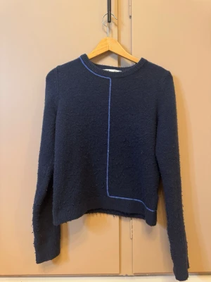 Mörkblå stickad tröja från Zara - Mörkblå stickad tröja från Zara med lång ärm och rund halsringning. Tröjan har en tunn ljusblå linje som detalj framtill och baktill, samt ribbade muddar vid ärmslut och nederkant. Perfekt för lager-på-lager under kyliga dagar.
