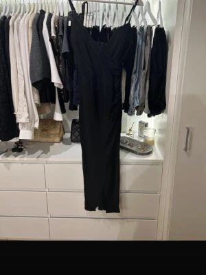 Svart glittrig långklänning - Säljer en svart långklänning med tunna axelband och omlott framtill. Klänningen har ett glittrigt tyg som ger en snygg effekt och dragkedja i ryggen. Perfekt för dig som vill sticka ut lite extra.