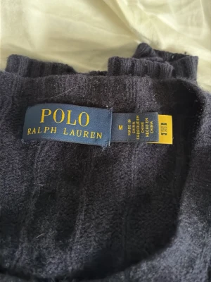 Mörkblå kabelstickad tröja Polo Ralph Lauren - Mörkblå Ralph lauren tröja som är stickad med en kashmir blandning, köpt på NK i Stockholm och sista bild som bevis att den är äkta! Hör av dig vid frågor! Inga deffekter alls och knappt använd 