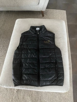 Svart dunväst från EA7 Emporio Armani - Snygg svart dunväst från EA7 Emporio Armani med quiltad design och dragkedja framtill. Västen har hög krage och diskret logga på bröstet. Perfekt att slänga över en hoodie eller tröja för en sportig look.