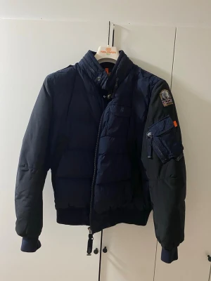 Mörkblå pufferjacka från Parajumpers - Säljer en mörkblå pufferjacka från Parajumpers med patch på ärmen, flera fickor och dragkedja framtill. Jackan har hög krage och ribbade muddar. Insidan är beige och vadderad för extra värme. Perfekt för kalla dagar.