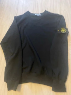 Svart sweatshirt från Stone Island - Säljer en svart sweatshirt från Stone Island med den klassiska loggan på vänster ärm. Tröjan har rund halsringning, ribbade muddar och är tillverkad i mjuk bomull. Perfekt för dig som gillar streetwear och vill ha en clean look.