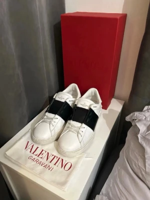Valentino Garavani vita sneakers - Säljer ett par snygga vita Valentino Garavani sneakers med bred svart rem över vristen. Skorna har rund tå, platt sula och klassisk logga på plösen. Tillverkade i skinn och levereras med originalpåse och röd box. Perfekta för dig som gillar exklusiv streetstyle. Dessa är helt nya och jag säljer dom då de inte passar mig, sjukt snygga, priset ej hugget i sten så kom med prisförslag!