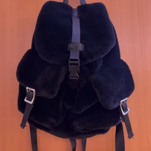 Svart ryggsäck i fuskpäls från Bik Bok - Mysig svart ryggsäck i fuskpäls från Bik Bok med spännen och justerbara remmar. Väskan har flera fickor och stängs med både spänne och dragsko. Perfekt för dig som vill ha en unik accessoar med mjuk känsla.