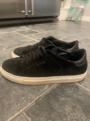 Svarta sneakers från Axel Arigato - Säljer ett par svarta sneakers från Axel Arigato med ovandel i mocka och detaljer i skinn. Skorna har klassisk låg profil, vita gummisulor och snörning framtill. Diskret Arigato-logga på sidan och bak på hälen. Perfekta till jeans eller chinos.