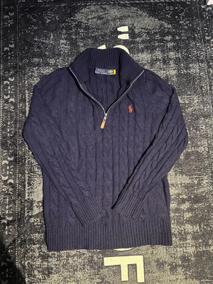 Marinblå stickad polotröja Polo Ralph Lauren - Säljer en marinblå stickad polotröja från Polo Ralph Lauren med klassiskt kabelstickat mönster och röd broderad logga på bröstet. Tröjan har half zip-dragkedja och ribbad krage, perfekt för kyliga dagar.