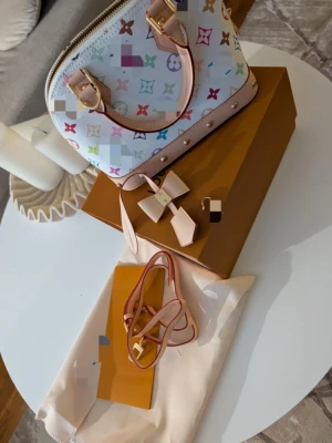 Vit handväska - Säljer en ikonisk vit handväska . Väskan har ljusrosa detaljer, guldfärgade metalldelar och medföljande axelrem samt söt rosettdetalj. Insidan är beige och väskan stängs med dragkedja.