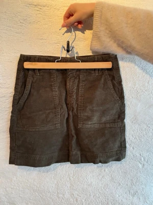 Mörkgrön manchesterkjol med fickor - Säljer en mörkgrön kortkjol i manchester med stora fickor både fram och bak. Kjolen har bälteshällor och en rak passform som ger en chill vibe. Perfekt att styla med stickad tröja eller boots för en snygg look.