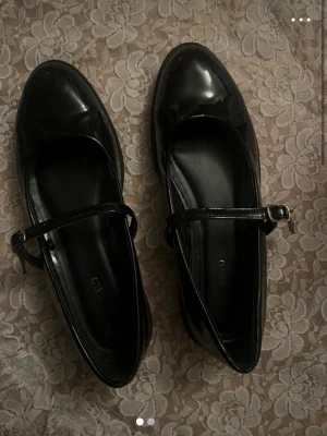 Svarta Mary Jane ballerinaskor - Säljer ett par svarta Mary Jane-inspirerade ballerinaskor i glansigt syntetmaterial. Skorna har rund tå, justerbart spänne över foten och en chunky platt sula. Perfekta för dig som vill ha en klassisk men edgy look.