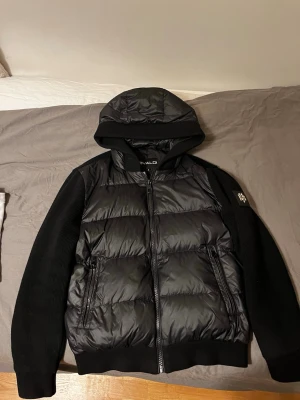 Svart puffer cardigan från Bvaldi - Säljer en svart puffer cardigan från Bvaldi i storlek S. Jackan har huva, ribbade stickade ärmar och quiltad kropp i syntetmaterial. Två dragkedjeförsedda fickor och snygg logga på ärmen. Perfekt för kyliga dagar. Den har en liten skada precis bredvid luvan men den är inte så synlig! Bara att höra av sig vid fler frågor