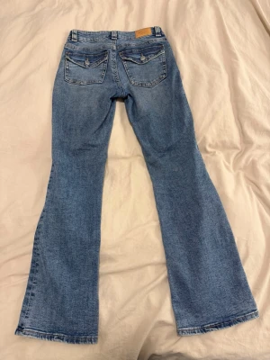 Blå bootcut jeans från Gina Tricot - Säljer ett par klassiska blå bootcut jeans från Gina Tricot. Jeansen har låg midja, fem fickor och snygga detaljer med knappar på bakfickorna. Tillverkade i mjukt denimtyg med lätt tvättad look och raka ben som går ut i en bootcut.