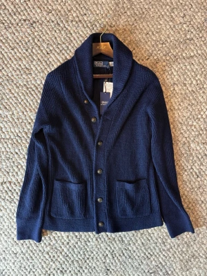 Ralph Lauren cardigan  - Säljer en marinblå stickad kofta från Polo Ralph Lauren. Koftan har grovstickad struktur, två framfickor, knappar hela vägen och en bred sjalkrage. Perfekt för lager-på-lager och chill höststil.