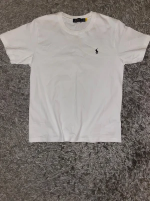  Polo Ralph Lauren - Klassisk vit t-shirt från Polo Ralph Lauren med rund hals och korta ärmar. Diskret broderad logga i mörkblått på bröstet. Tillverkad i mjuk bomull för en skön och clean look. Perfekt till jeans eller shorts för en enkel och stilren outfit.