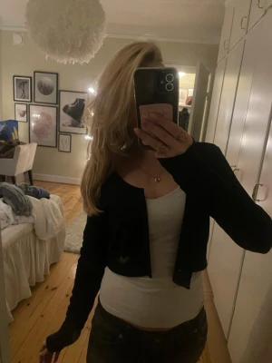 Svart croppad kofta med knappar - En svart, croppad kofta med långa ärmar och knäppning framtill. Koftan har en enkel och stilren design som passar perfekt till många olika outfits. Materialet känns mjukt och stretchigt, vilket gör den bekväm att bära.