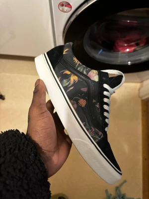 Svarta Vans sneakers med blommigt mönster - Säljer ett par Vans sneakers i svart med blommigt mönster på sidorna. Skorna har vit sula, vita snören och klassisk Vans-design. Ovandelen är i textil och mocka, vilket ger en snygg kontrast till det färgglada blommönstret. Perfekta för dig som vill sticka ut med din stil.