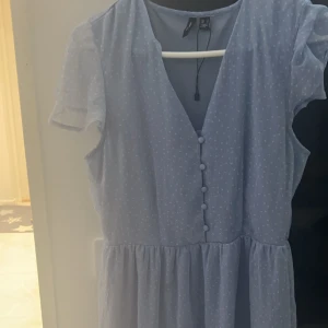 Ljusblå klänning med knappar - En ljusblå klänning med små vita prickar. Klänningen har korta ärmar och knappar framtill. Den har en v-ringad hals och en volangdetalj vid fållen.
