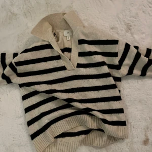 Randig stickad tröja från H&M - En bekväm och stilren stickad tröja från H&M med svarta och vita ränder. Tröjan har en krage och långa ärmar med ribbstickade muddar vid ärmslut och nederkant.