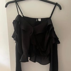 Svart blus från H&M - Säljer denna eleganta svarta blusen från h&m med volangdetaljer och off-shoulder design. Blusen har långa ärmar och smala axelband. Endast använd en gång. 🖤✨🌸