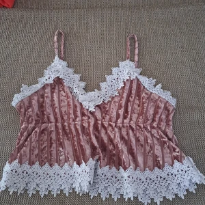 Rosa sammetstopp med spetsdetaljer - En rosa sammetstopp med vita spetsdetaljer runt halsen, ärmhålen och nederkanten. Toppen har smala axelband och en v-ringad halslinje. Den är ribbad och har en lös passform. det är pijamas top. Jag säljer den för att det är inte längre min storlek.