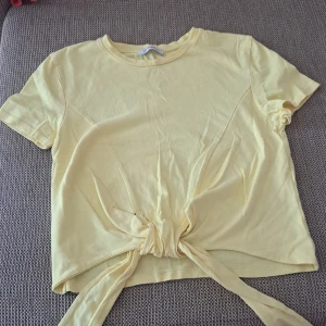 Gul kortärmad topp med knytning - En gul kortärmad topp med rund halsringning och knytning framtill. Toppen är tillverkad i ett mjukt och bekvämt material.