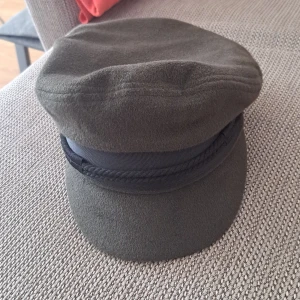 Grön keps med svart band - En grön keps med ett svart band och dekorativt rep. Kepsen har en mjuk form och är tillverkad i ett tygmaterial.