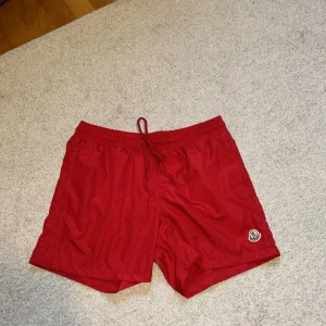 Röda Moncler badshorts  - Ett par röda Moncler badshorts med elastisk midja och dragsko. Shortsen har en liten Moncler-logga på vänster ben och är tillverkade i ett lätt och bekvämt material. Har endast blivit använda 3-4 gånger. 