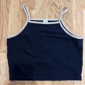 Svart linne från Lindex - Ett svart linne från Lindex med beige kant runt halsen och ärmhålen. Linnet har smala axelband och är tillverkat i Turkiet.