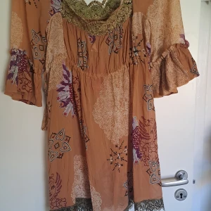 Annie Oakley Salon klänning - En vacker klänning från Annie Oakley i färgen mörk puder. Klänningen har ett mönster med blommor och geometriska former i olika färger. Den har långa ärmar med volanger och spetsdetaljer vid halsen.