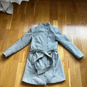 Kappa  - Stilren grå kappa i ull. Nyligen lagad dragkedja. Formsydd med vadderade axlar, bälte vid midjan.  Nypris 899kr