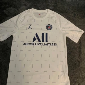 Vit fotbollströja från Jordan - En vit fotbollströja från Jordan med Paris Saint-Germain-logotypen på bröstet. Den har korta ärmar och ett mönster med ordet 'PARIS' över hela tröjan.