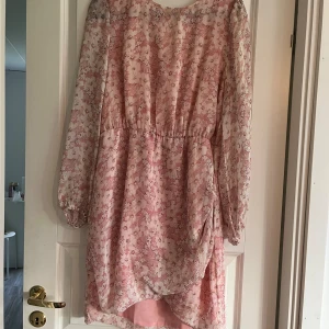 Blommig klänning - En blommig klänning med långärmade ärmar. Klänningen har en rosa och vit färgkombination med ett blommönster. Den har en lös passform och är tillverkad i ett lätt material. Aaknas en knapp på baksidan, men inga problem för den som kan sy