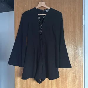 Svart klänning med knytning - En elegant svart shortsklänning med lång ärm och knytning framtill. Klänningen har en enkel och stilren design. Blixtlås i rygg. 