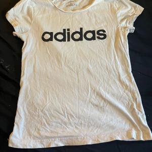 Vit t-shirt från Adidas - En vit t-shirt från Adidas med korta ärmar och en rund halsringning. T-shirten har en svart Adidas-logotyp tryckt på framsidan.