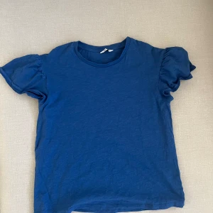 Blå t-shirt  - En nästan oanvänd mörkblå t-shirt med volang i armarna. Bra skick och bra passform. Mycket fin färg. Väldigt härlig och funkar året om